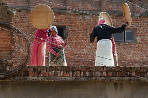 Sul tetto di un edificio alcune donne Newari, impugnando un grande disco in tessuto vegetale intrecciato da loro stesse, lo sventolano ritmicamente sul grano in modo da separare la crusca rispetto ai semi (essendo più leggera, vola via facilmente): questo movimento lo ripetono un numero infinito di volte, sino a quando quasi tutta la crusca è volata via, villaggio di Khokanà, Nepal 2018.