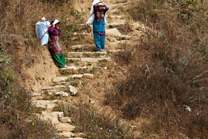 Due donne Newari si fermano stremate dopo aver trasportato sulle spalle un sacco enorme (contiene il raccolto) su di una scalinata di pietra ripida che collega i campi coltivati al vicino villaggio di Khokanà: li segue a breve distanza un uomo che, libero da ogni peso, sale dietro di loro con le braccia dietro la schiena. Nepal 2018