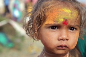 Anche i bambini partecipano alla Festa in onore della Dea Yallamma: anche loro sono ricoperti della polvere ocra di curcuma e rossa di kum kum, tra nuvole gialle e rosse che si addensano nell'aria ad ogni lancio da parte dei pellegrini festanti. Questa bimba dallo sguardo intenso e quasi imbronciato si chiama "Kinari" (tradotto in italiano, significa "riva"). Dintorni del villaggio di Saundatti, Regione del Karnataka, India 2015.