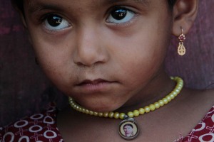 Primo piano di Chandraki, bimba Hindu di cinque anni orfana della mamma: nel medaglione posto al centro della collana porta l'immagine della defunta. Il suo sguardo carico di dolore e tristezza testimonia che il cammino della sua vita è in salita. Villaggio di Saundatti, Regione del Karnataka, India 2015.