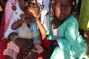 Bambina Haoussa nasconde il viso con una bottiglia d'acqua mentre è tra le braccia della sua mamma, con accanto la sorellina sorpresa e quasi intimorita alla vista della mia macchina fotografica, Festival dell'Air, Iferouane, Deserto del Ténéré, Regione dell'Air, Niger 2020