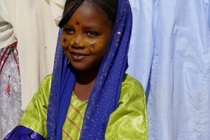 Bambina Tuareg Kel Ferouane con i suoi vestiti tradizionali eleganti e sobri: una tunica pregevole (tabanik iloumar), un raffinato velo sul capo che lascia scoperto il viso (iseggelmas alecho), presenta sul viso graziosi disegni realizzati con pigmenti organici naturali, sulle mani e su parte delle braccia esibisce dei tatuaggi temporanei eseguiti con una miscela di hennè, hargus ed acqua. Festival dell'Air, Oasi di Iferouane, Deserto del Ténéré, Niger 2020